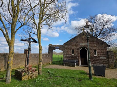 Bakstenen kapel met kruisbeeld en lantaarnpaal, omgeven door bomen en gras, onder blauwe lucht.