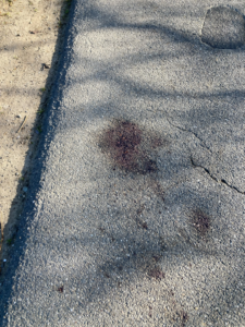 Gebarsten asfalt trottoir met donkere vlekken en zandstrook links. Schaduwen zichtbaar.