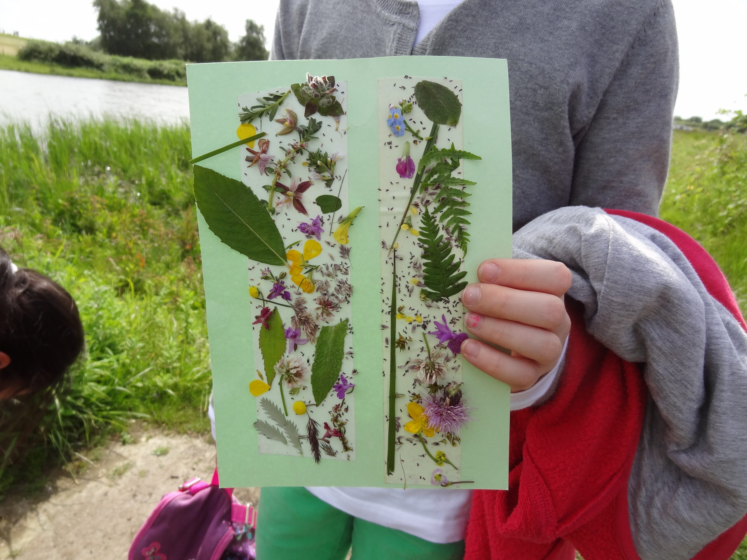 Een kind houdt een vel gekleurd papier vast met geplakte bloemen langs een meer.