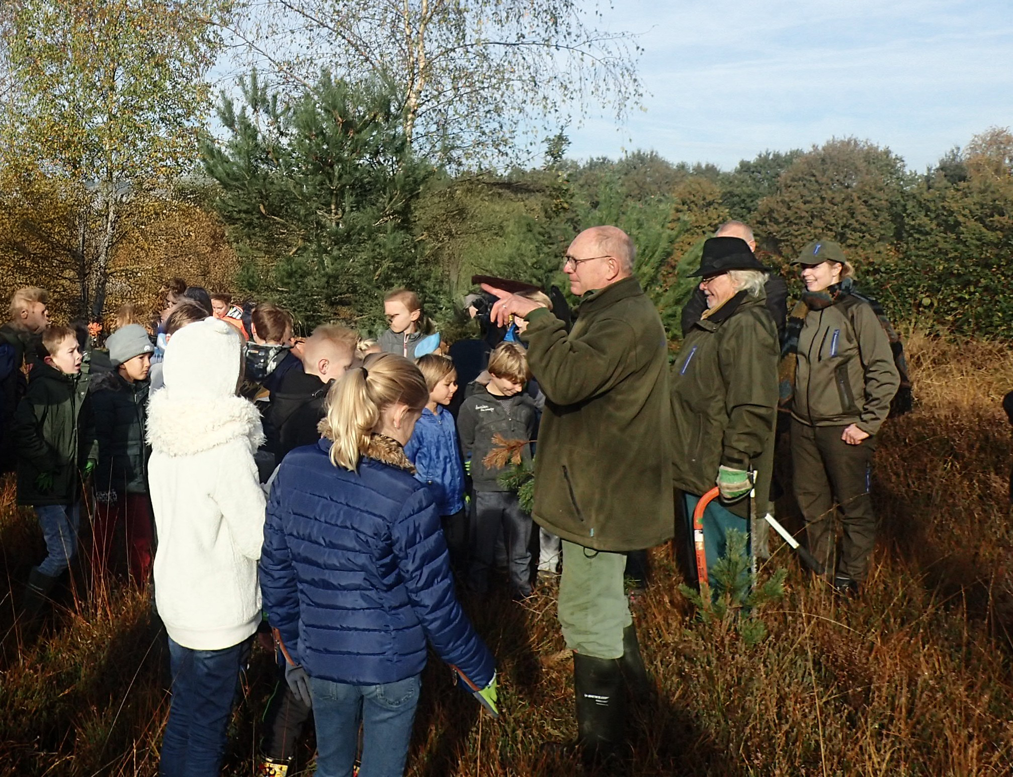 Een groep kinderen krijgt uitleg van volwassenen in een bosrijke omgeving.