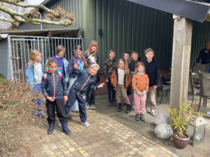 Groep kinderen poseert buiten, met een metalen hek en een groene muur op de achtergrond.