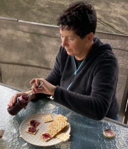 Persoon smeert jam op crackers aan een glazen tafel buiten.