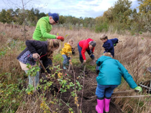 Enkele volwassenen en kinderen planten samen bomen in een veld met hoge grassen.