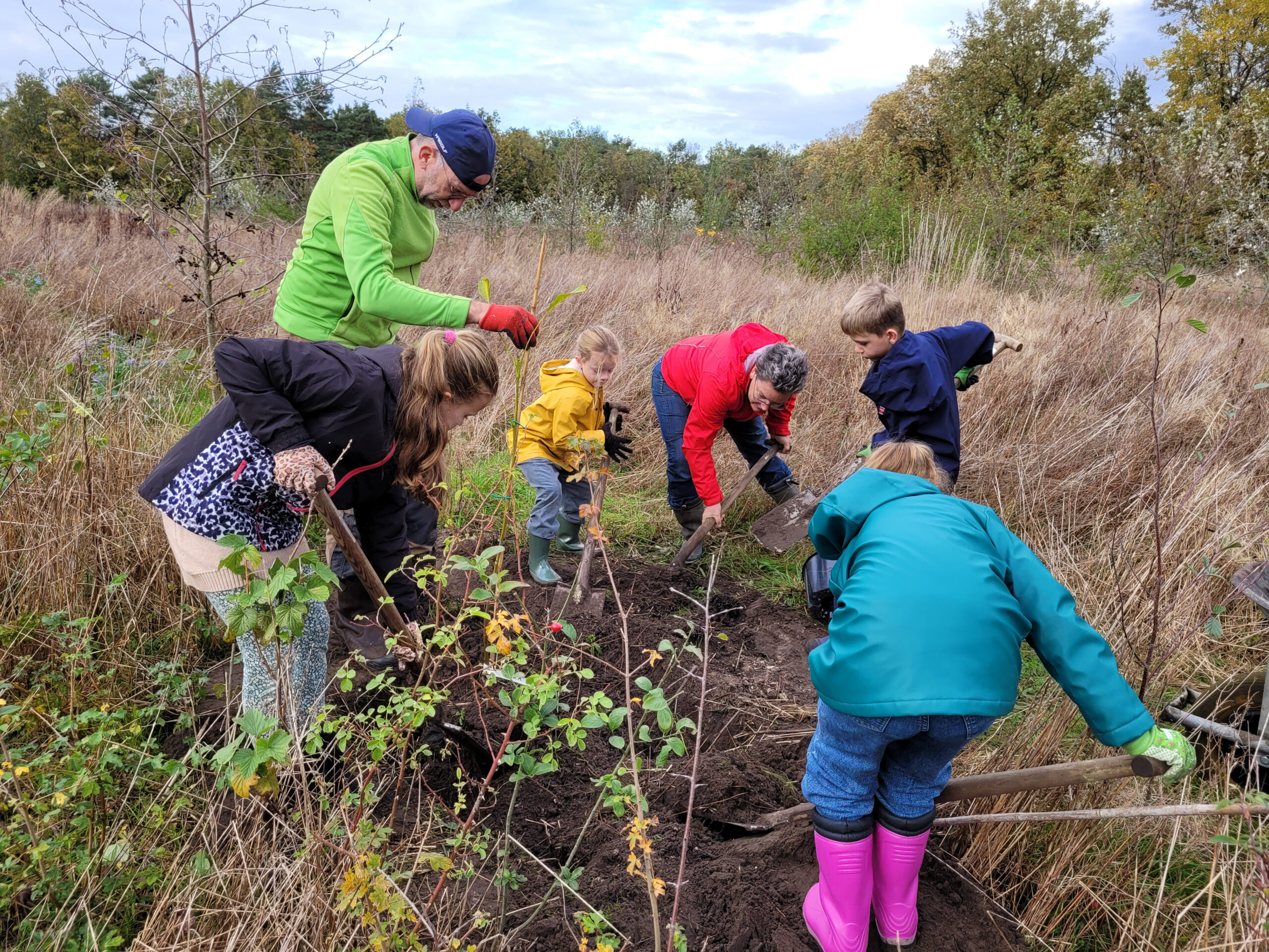 Enkele volwassenen en kinderen planten samen bomen in een veld met hoge grassen.