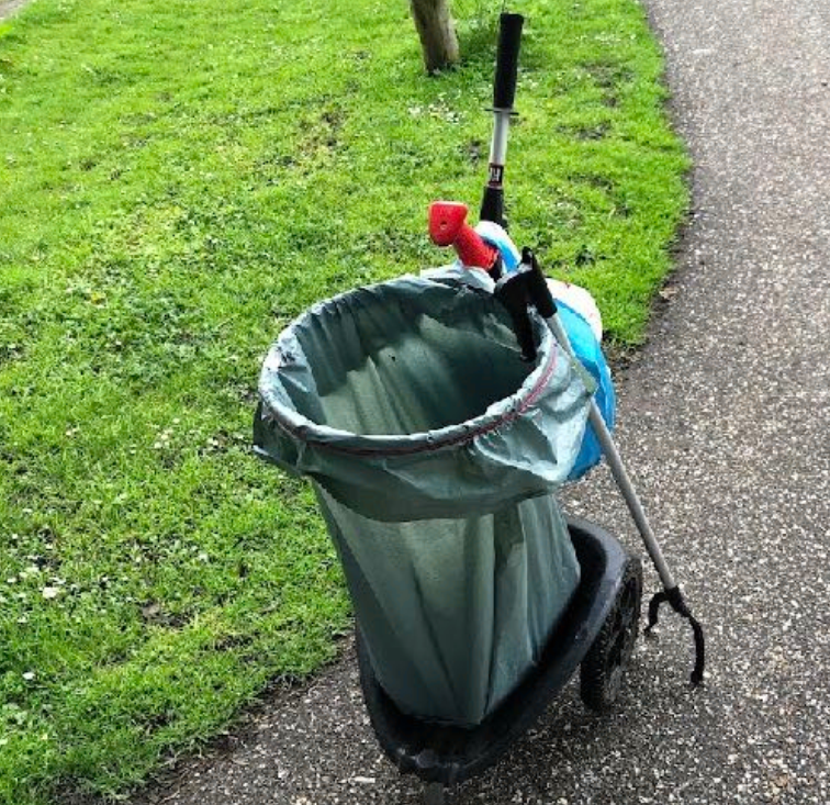 Afvalcontainer met grijpstok en rode handpop ernaast op trottoir naast grasveld.