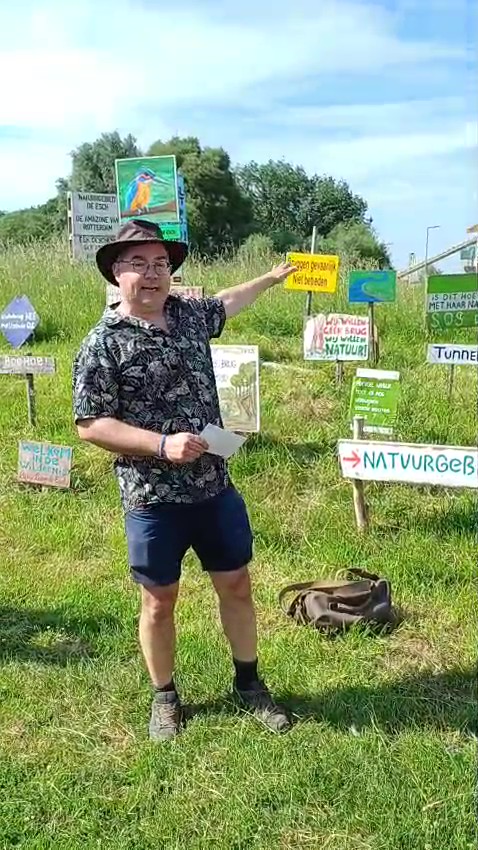 Man in korte broek en hoed wijst naar protestborden in een groen veld.