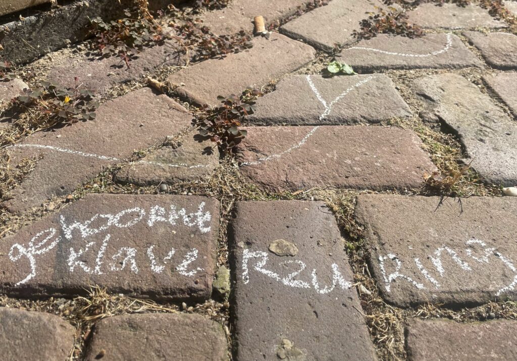 Stoeptegel met krijttekst en tekeningen, gras en onkruid zichtbaar tussen de voegen.