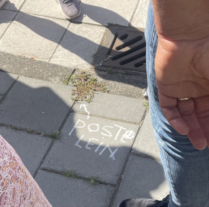 Voetgangers op stoep met krijttekening "ROST PLEIN" en pijl naar afvoerputje.