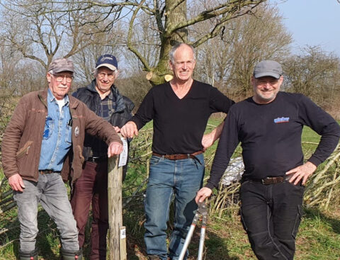 Vier mannen poseren buiten naast een houten paal en een boom op een zonnige dag.