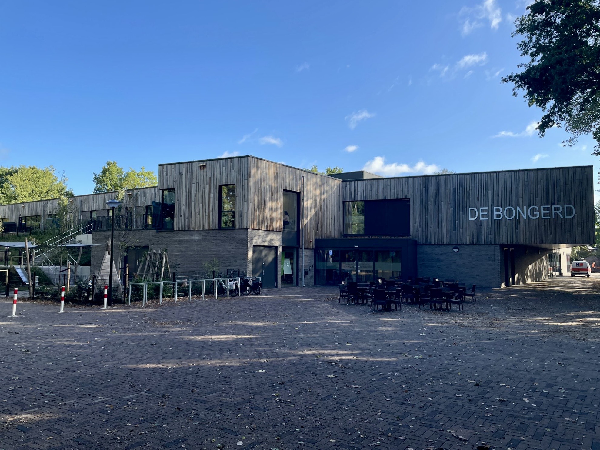 Modern gebouw met houten gevel en glazen ramen, naam "DE BONGERD" zichtbaar. Aangelegd plein ervoor.