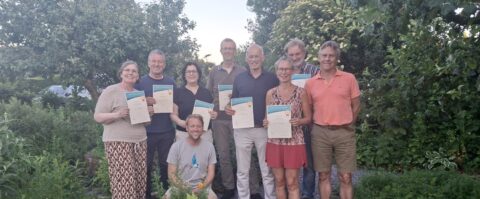 Groep mensen poseert buiten met certificaten, omringd door groen en bomen.