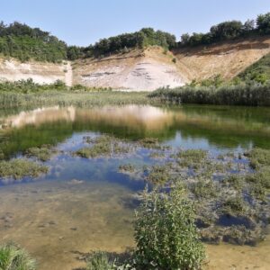 Vijver met helder water en groen, omringd door heuvels en begroeiing onder een blauwe hemel.