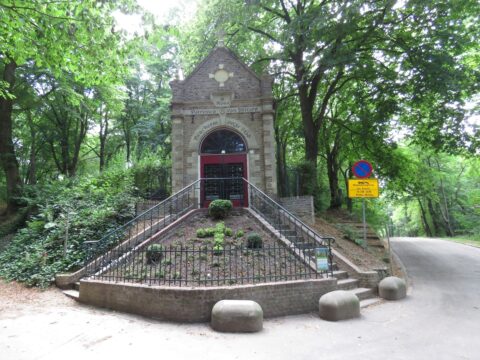 Kapel in bos, omringd door bomen, trap en hekwerk ervoor. Verkeersbord naast weg.