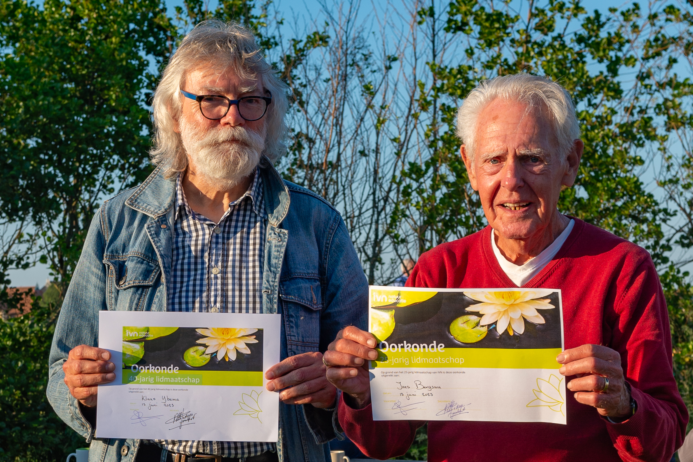 Twee mannen tonen certificaten voor 40-jarig lidmaatschap, buiten bij een zonnige achtergrond.