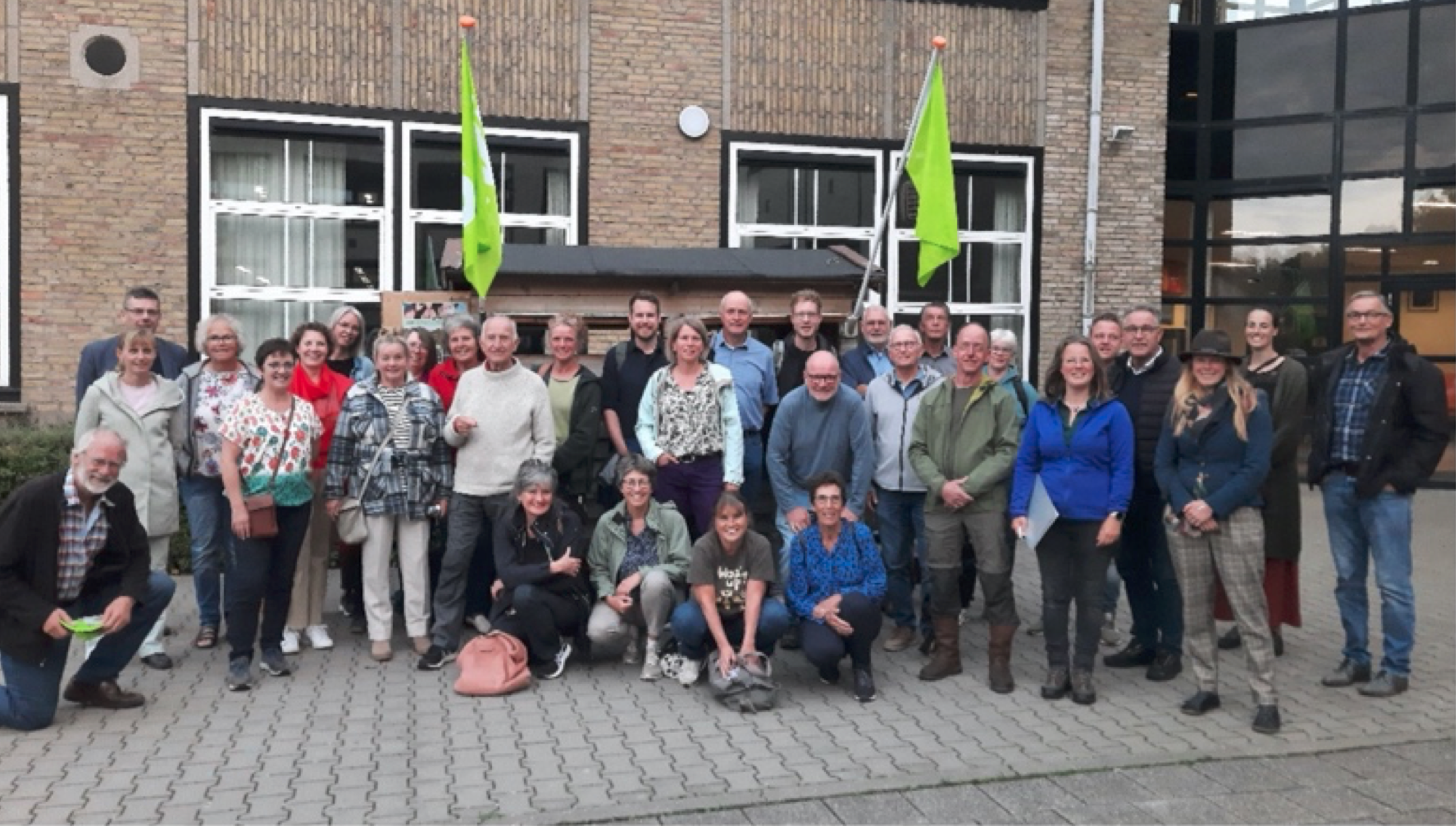 Groep mensen poseert voor een gebouw met groene vlaggen op de achtergrond.
