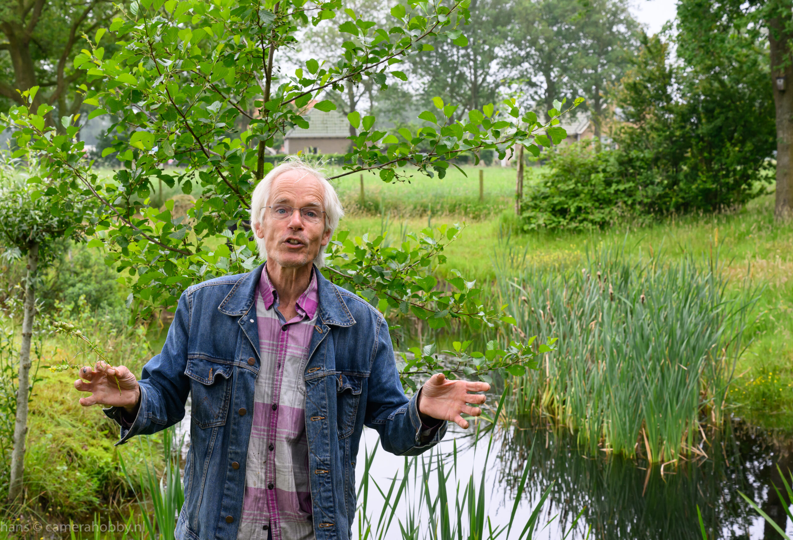 Een oudere man in een tuin met vijver en groen, draagt een spijkerjasje en praat buiten.