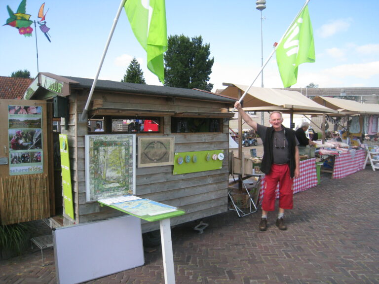 Man met groene vlag bij houten marktkraam, versierd met kaarten en posters, op een markt met kraampjes.