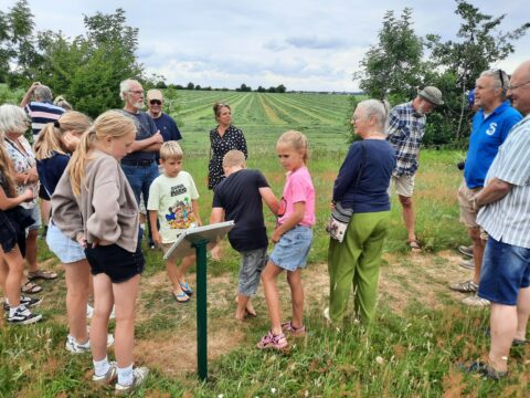 Groep mensen leest een bord naast een veld met gewassen, sommigen kijken nieuwsgierig omhoog.