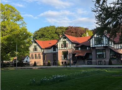 Groot, klassiek gebouw met rood dak in groene omgeving onder blauwe lucht.