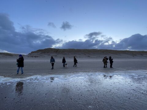Mensen wandelen bij zonsondergang op een verlaten strand met duinen en bewolkte lucht.