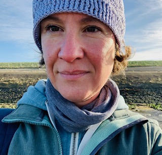 Persoon met gebreide muts en sjaal staat buiten op steenachtige ondergrond.