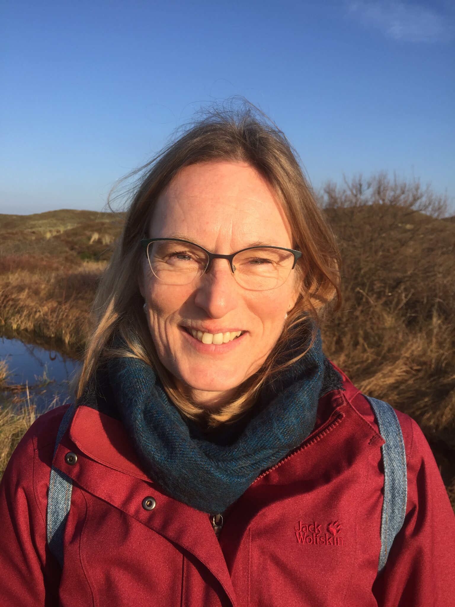 Vrouw in rode jas glimlacht in zonnig, grasrijk landschap met blauwe lucht.