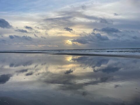 Strand bij zonsondergang, wolken en zon reflecteren op het natte zand.