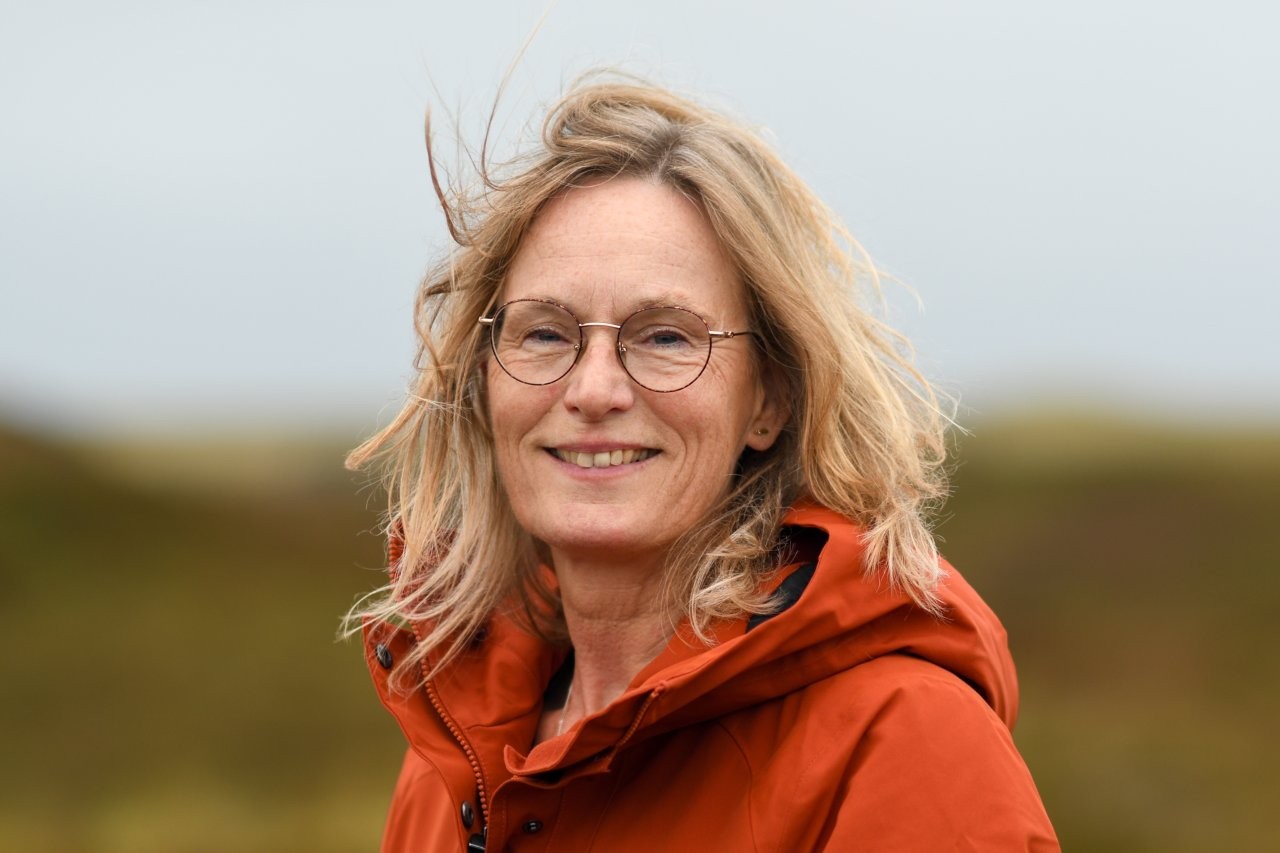 Vrouw met bril in oranje jas buiten, glimlachend met winderig haar tegen een wazige achtergrond.