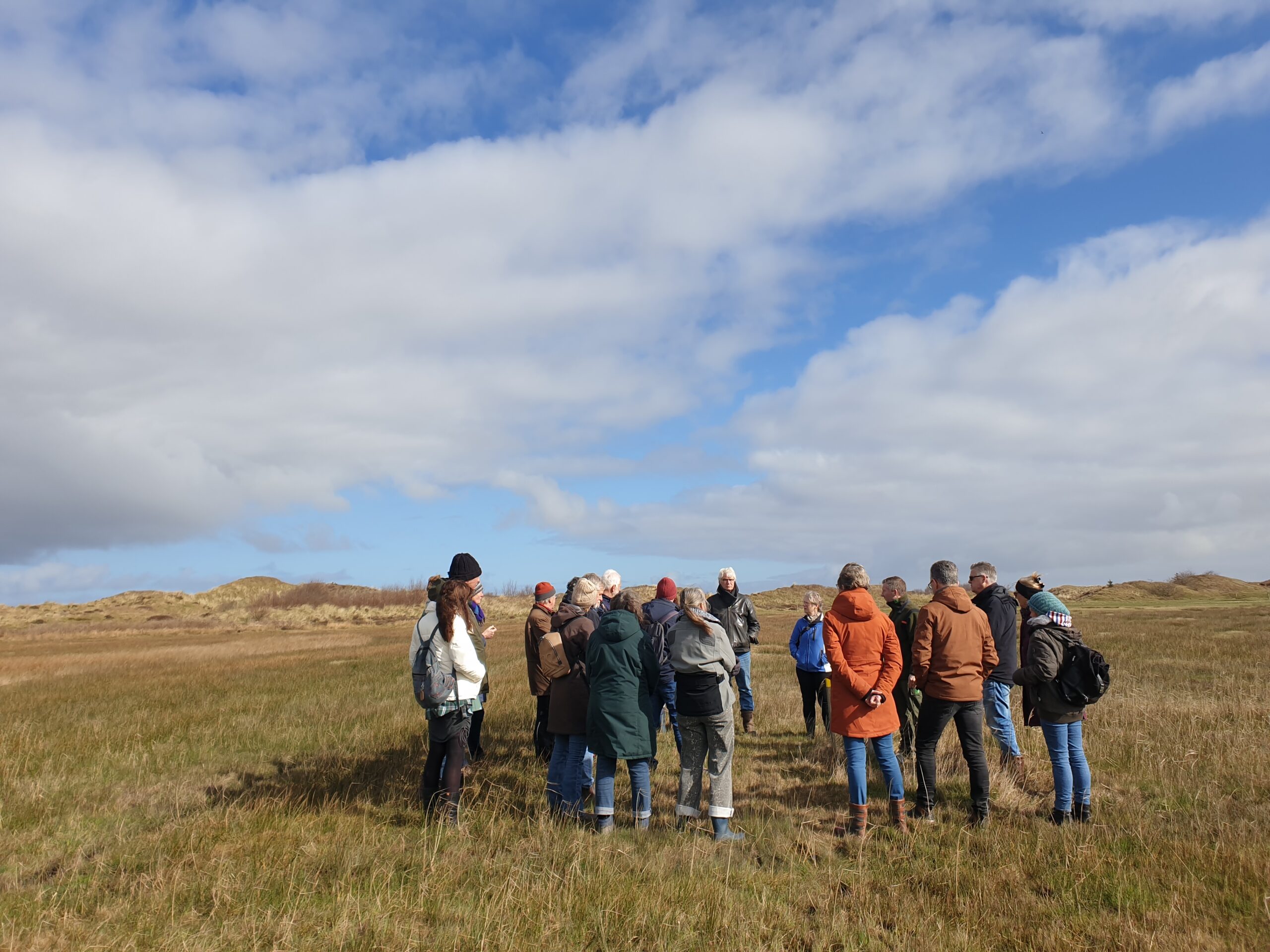 Groep mensen in winterkleding, buiten op een grasland onder een bewolkte blauwe hemel.