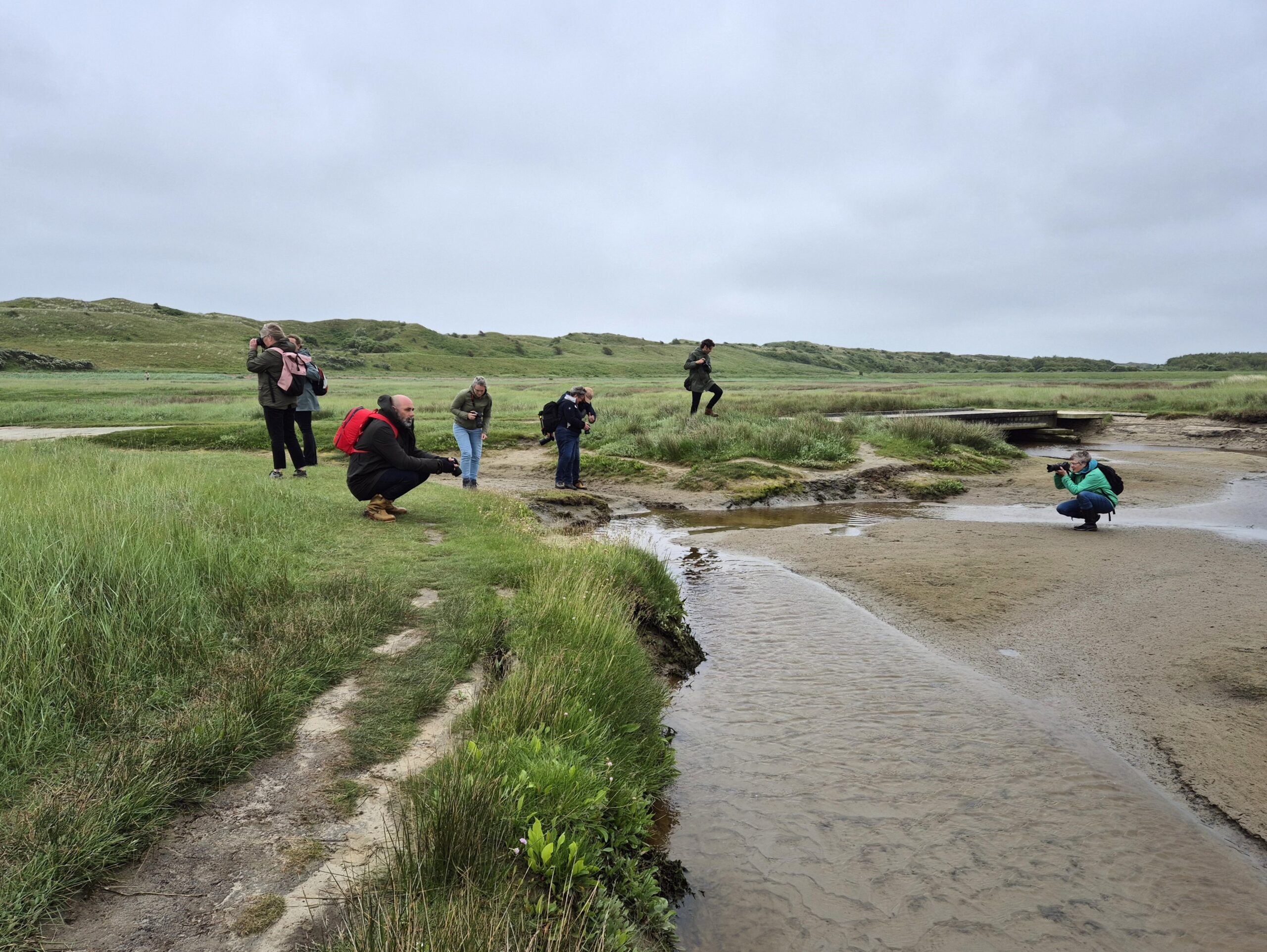 Mensen fotograferen een riviertje in een weids, groen landschap onder een bewolkte hemel.