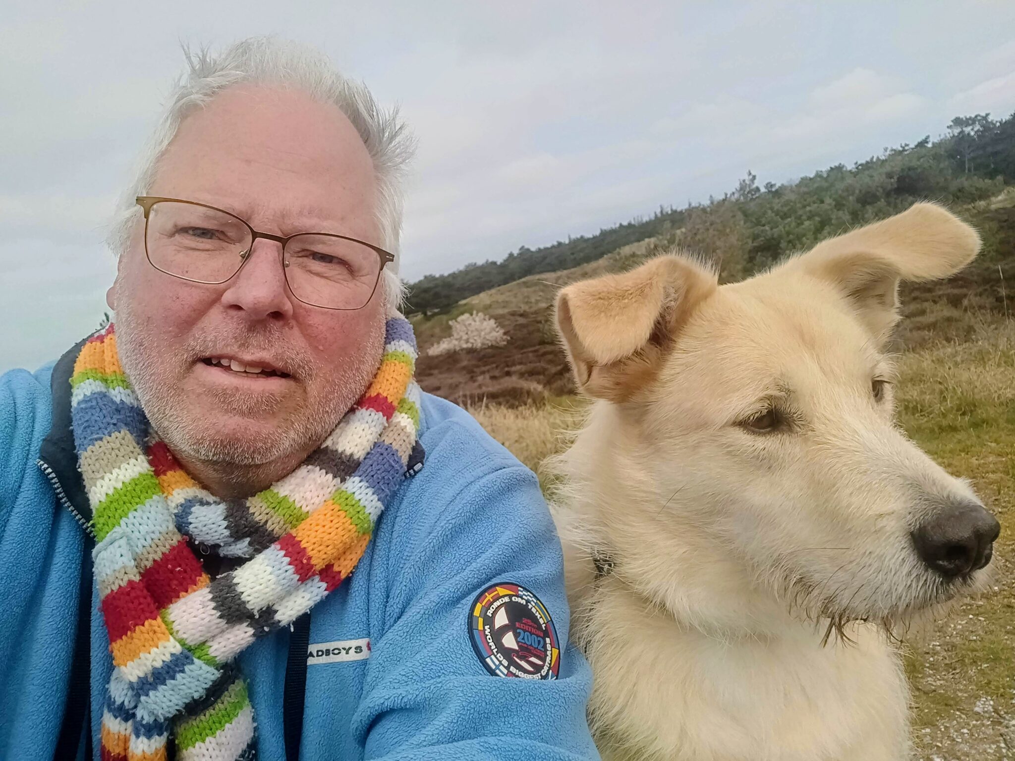 Man met gekleurde sjaal en hond in natuurgebied, bewolkte lucht en groene achtergrond.