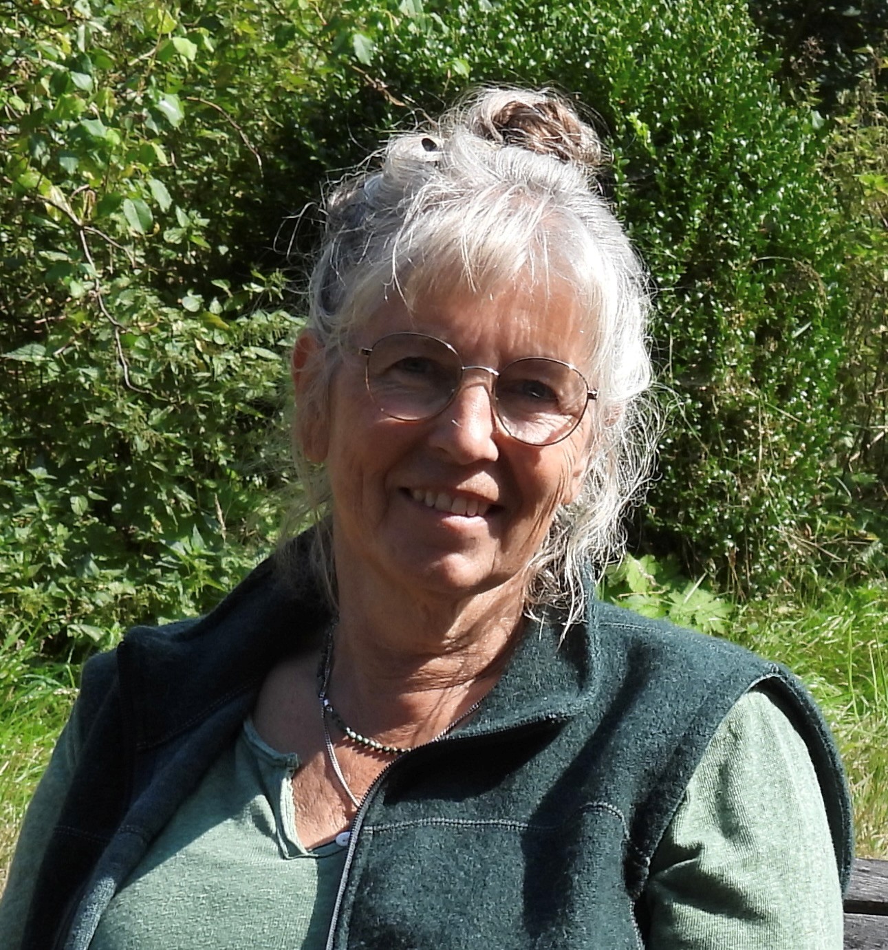 Oudere vrouw met bril glimlacht buiten in een groene omgeving.