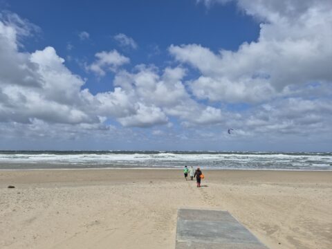 Strand met bewolkte lucht, ruwe zee en drie mensen die naar het water lopen.