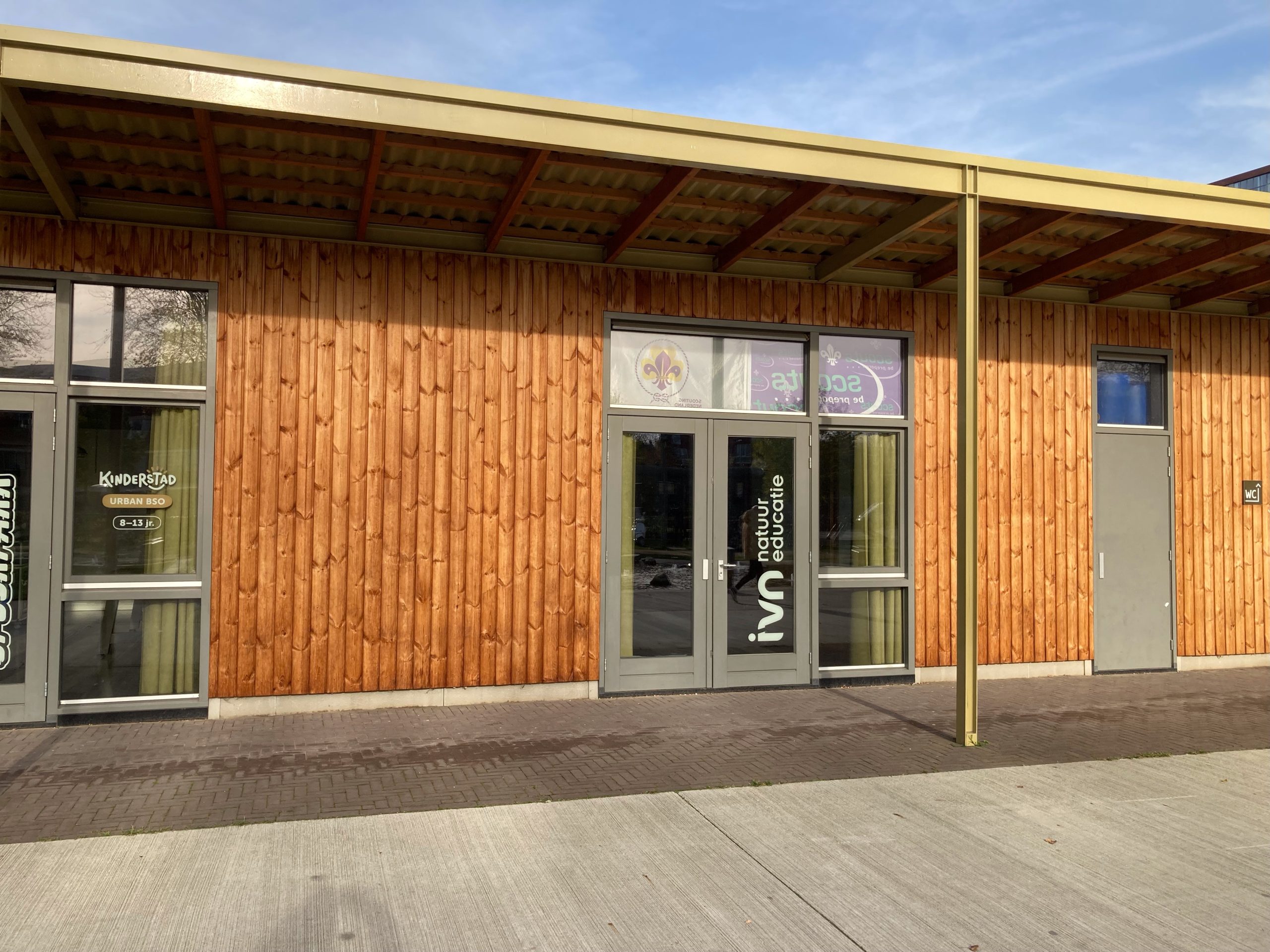 Houten gebouw met ramen, deuren en logo's van Kinderstad en IVN Natuureducatie op de gevel.
