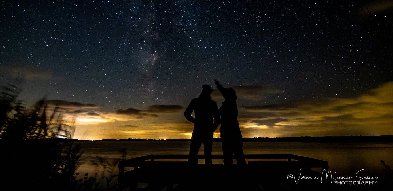 Twee silhouetten kijken naar sterrenhemel boven verlicht landschap en meer.
