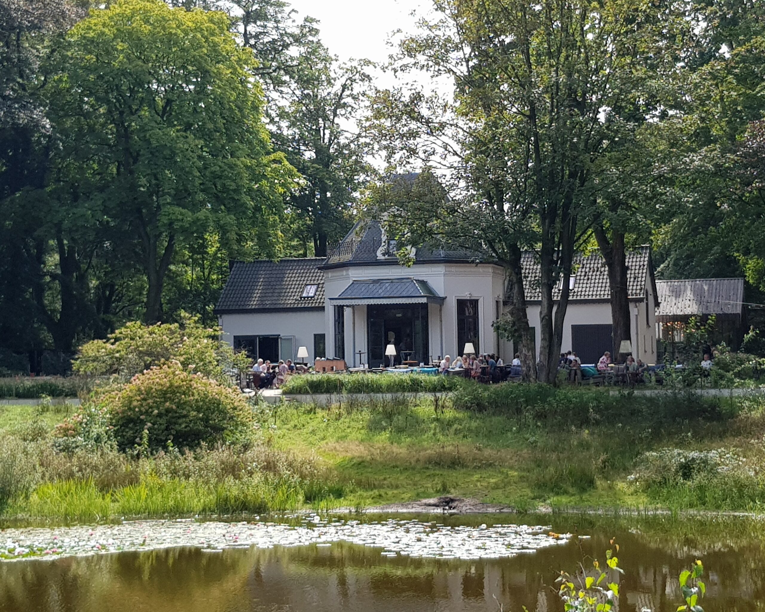 Een paviljoen met terras zit vol mensen, omringd door een vijver en groene bomen.