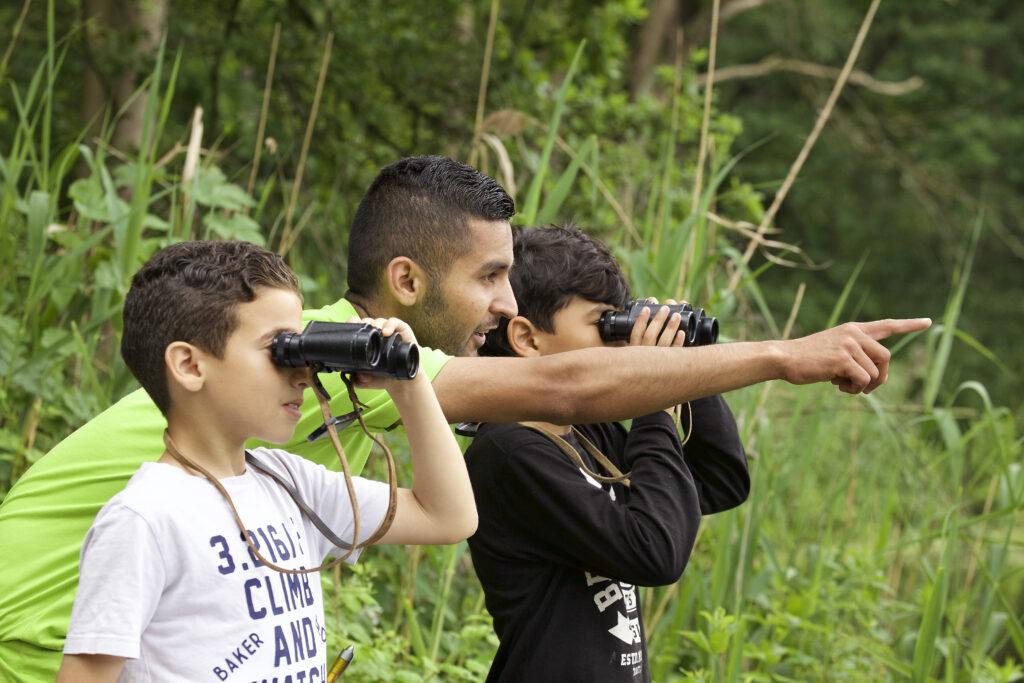 Twee kinderen en een volwassene vogels aan het spotten met verrekijkers in een groen natuurgebied.