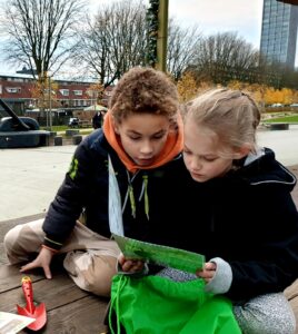 Twee kinderen lezen samen een kaart in een park, met een groene tas en schepje naast hen.