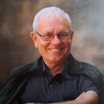 Een oudere man met bril lacht en draagt een donker shirt en jas over de schouder.