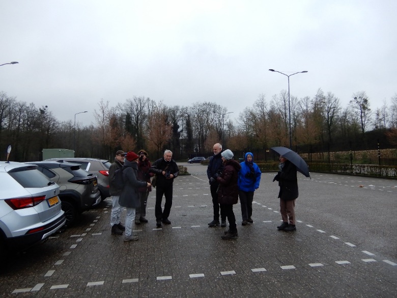 Groep mensen staat op een parkeerplaats in een park, onder een bewolkte hemel met enkele paraplu's.