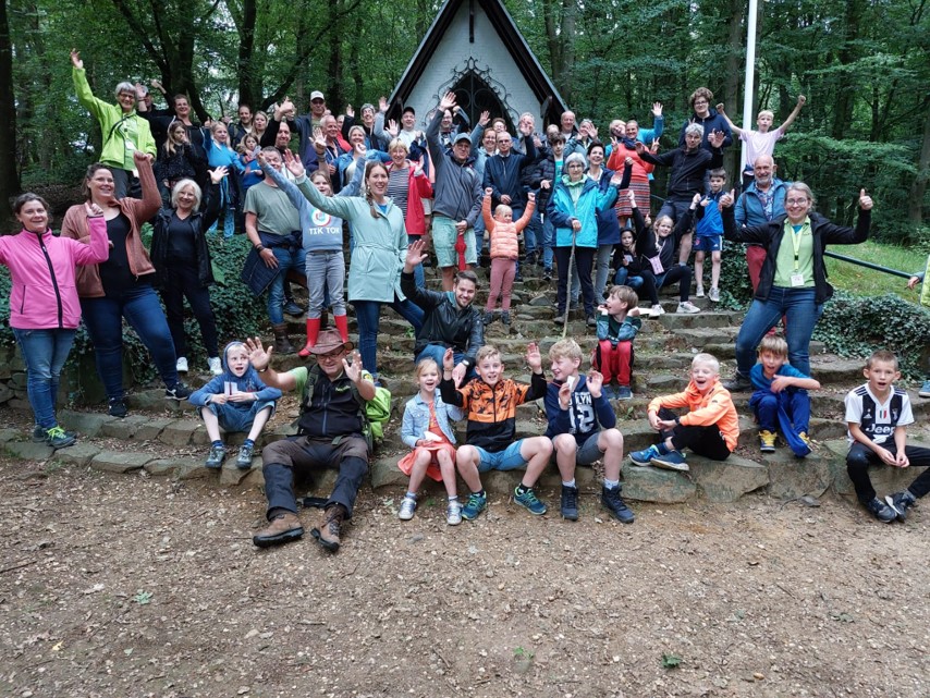 Groep mensen poseert vrolijk voor een kapel in een bosrijke omgeving.