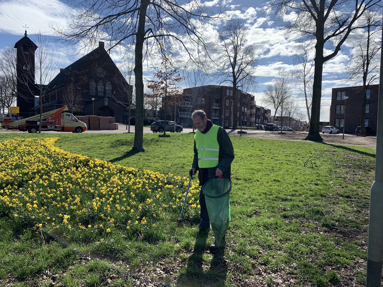 Man met veiligheidsvest verzamelt afval bij bloemenveld in park, kerk en gebouwen op achtergrond.