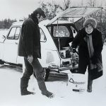 Twee mensen bij een auto in een besneeuwd landschap, bezig met spullen uitladen.