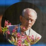 Oude man met bril kijkt naar roze bloemen in een tuin.