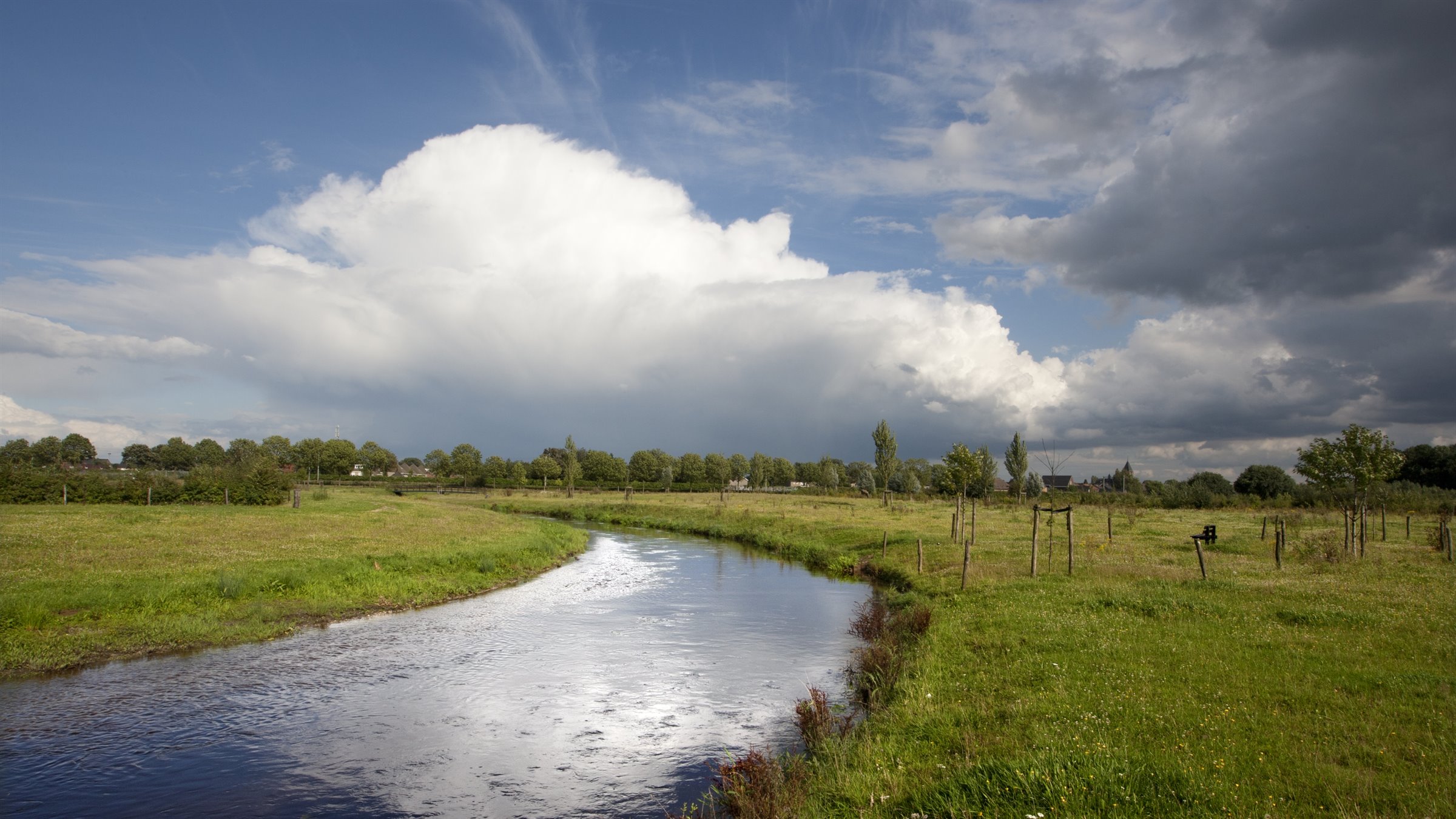 Landschap met een kronkelende rivier, groene weiden en grote, witte wolken onder een blauwe lucht.