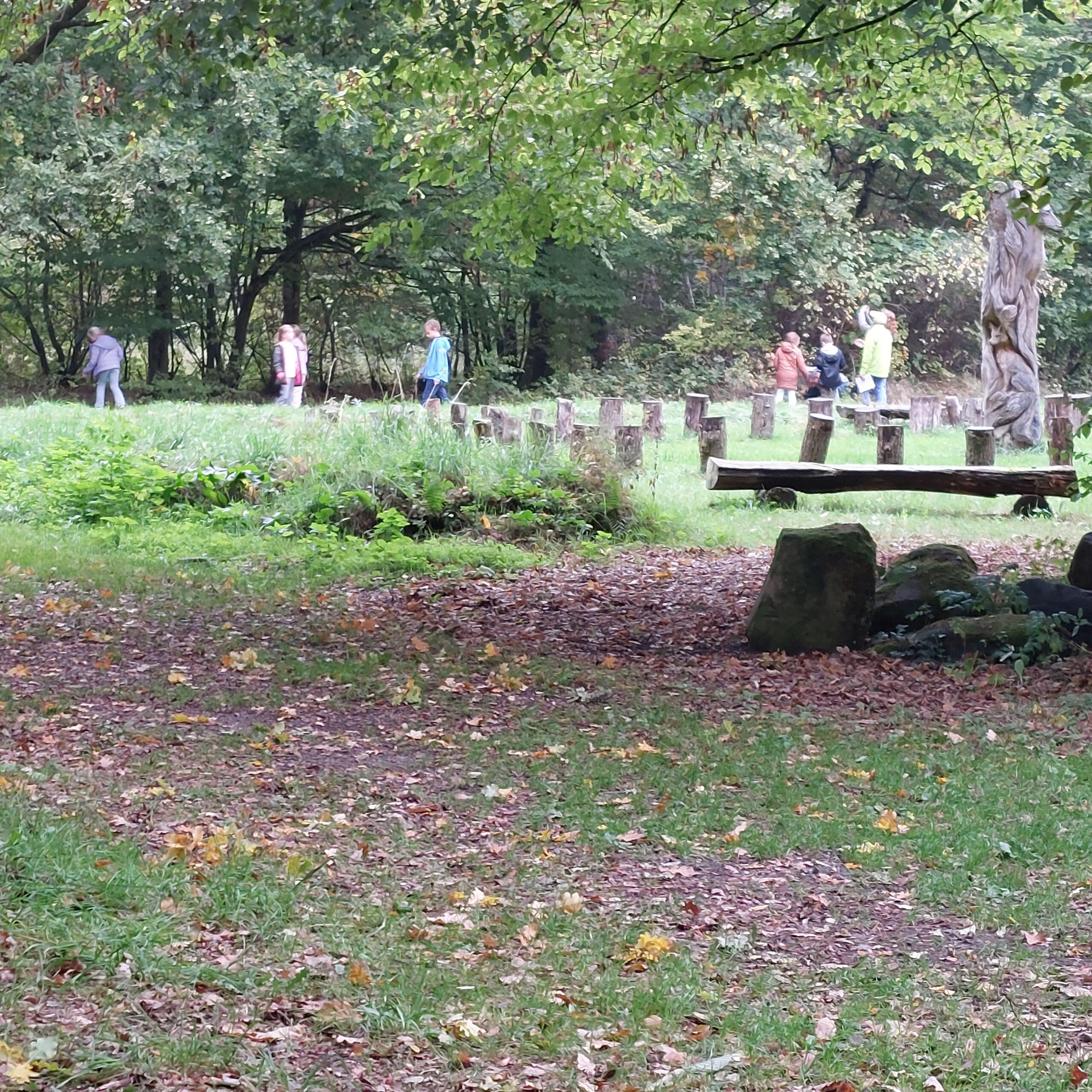 Mensen wandelen in een bos met een houten sculptuur en boomstammen op de achtergrond.