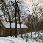 Bakstenen gebouw in besneeuwd bos, met kale bomen en hekwerk.