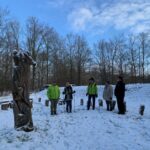 Vijf mensen staan bij een houten sculptuur in een besneeuwd, bebost gebied.