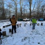 Twee personen in winterkleding staan ​​in een besneeuwd park met geparkeerde auto's en bomen.