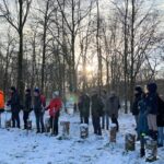 Groep mensen in winterkleding staat in besneeuwd bos met laagstaande zon erachter.